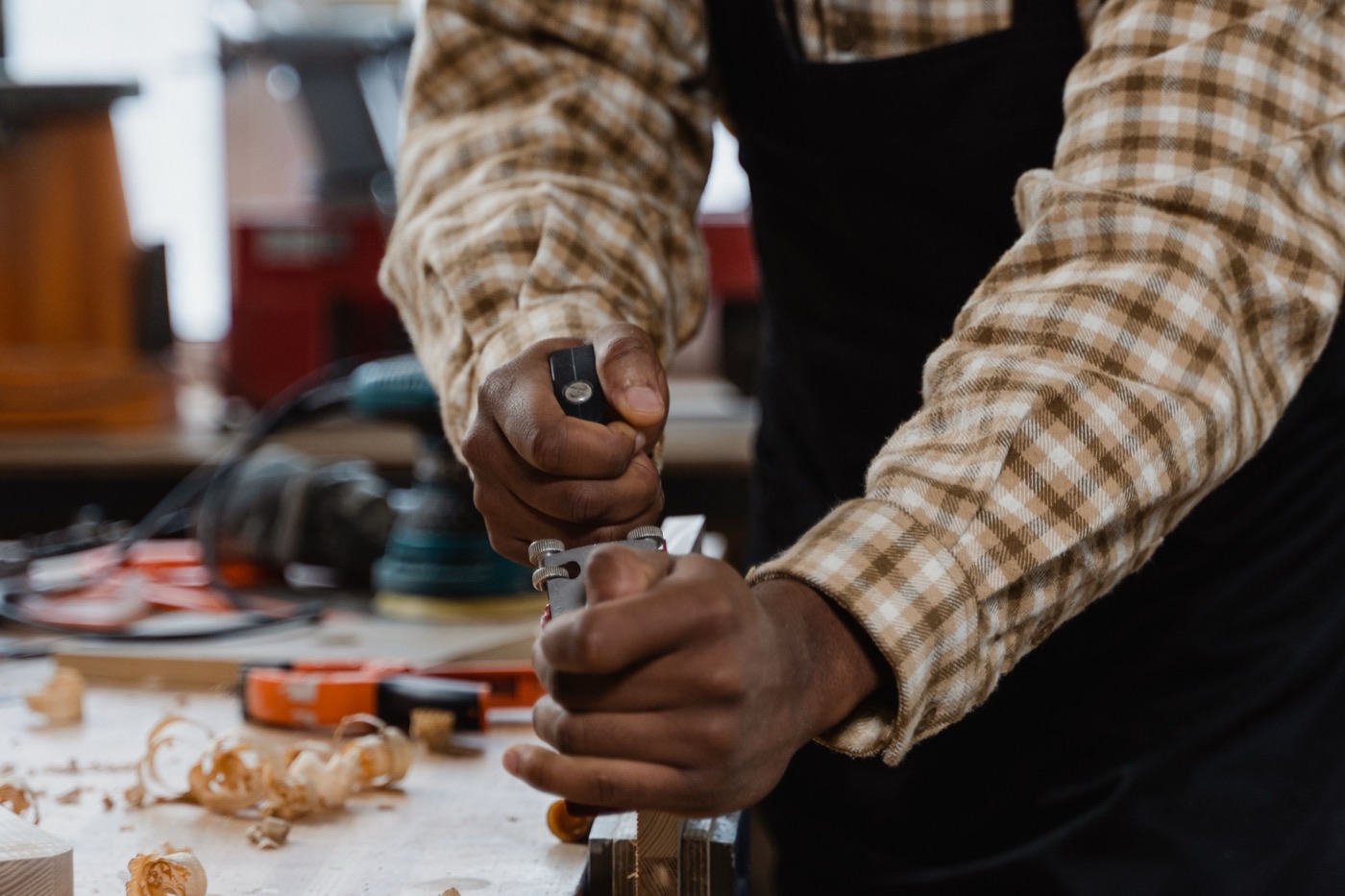 Close-up of a woodworker in a plaid flannel shirt and dark apron gripping a small hand plane at a workbench scattered with wood shavings.