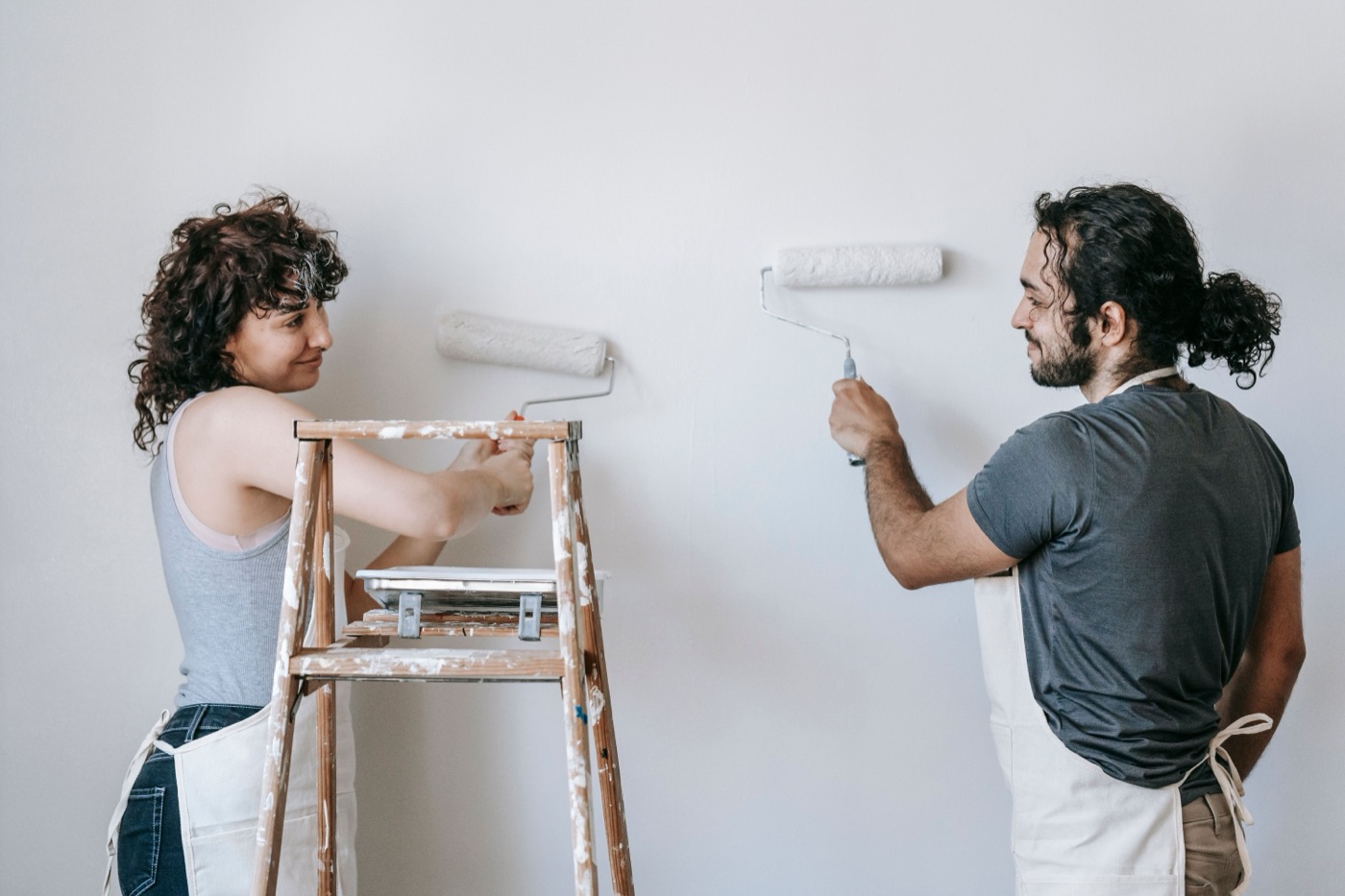 A smiling couple in canvas aprons roll white paint onto a wall together, one leaning on a paint-splattered wooden stepladder.