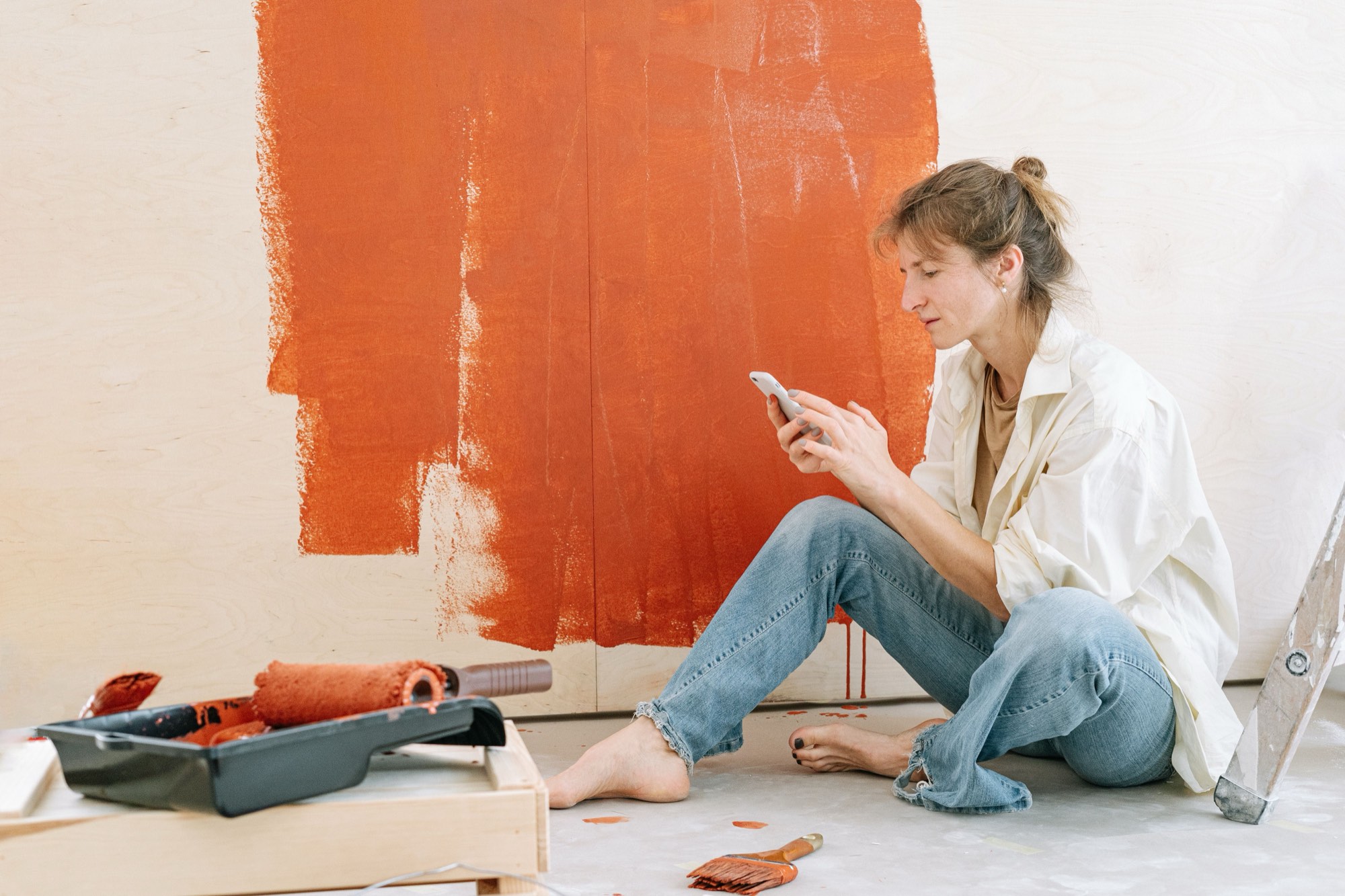 A woman in a cream work shirt and jeans sits barefoot on a drop cloth mid-way through painting a wall burnt orange, checking her smartphone, with a paint roller and tray beside her.