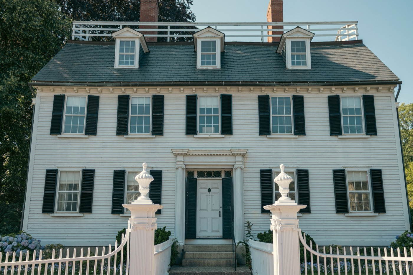 A white clapboard Georgian colonial house with black shutters, three dormers, twin brick chimneys and a white picket fence with urn-topped gateposts under a blue sky.
