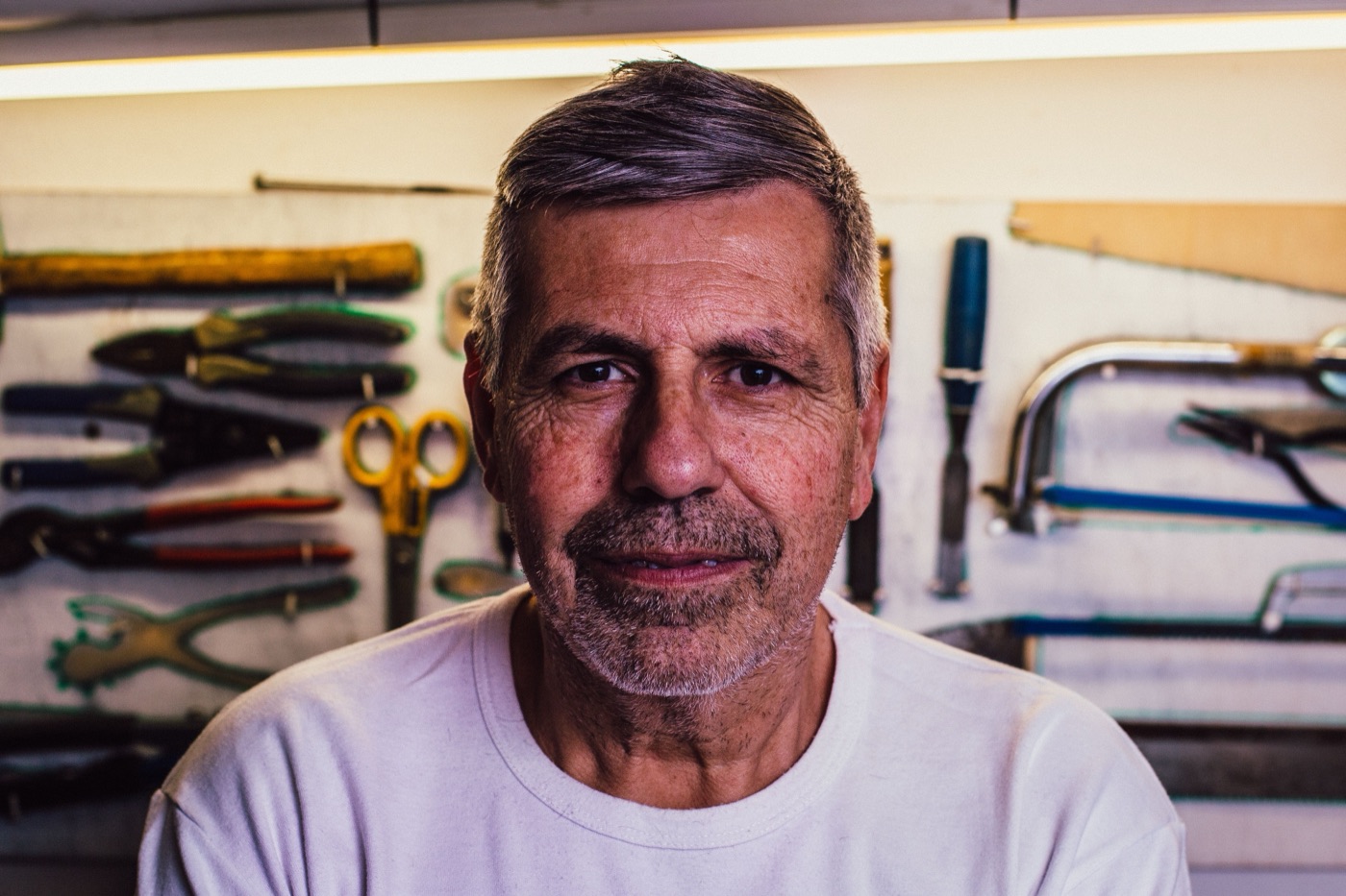 Portrait of a gray-haired older man in a white T-shirt looking into the camera, with pliers, a hacksaw and hand tools hanging on the pegboard behind him.
