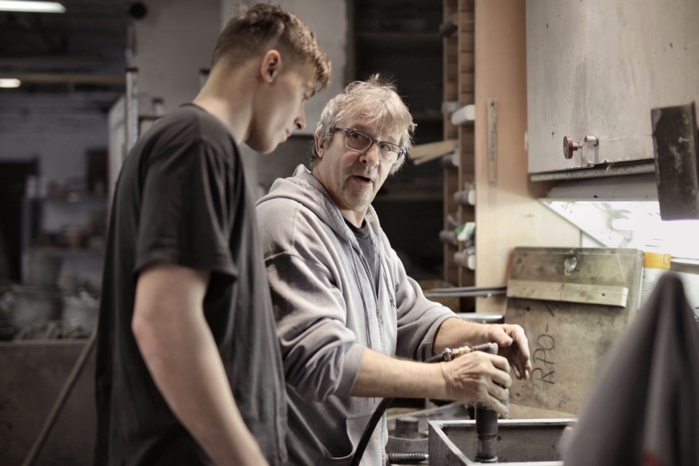 An older craftsman in glasses and a gray hoodie explains a technique to a young apprentice while working at a bench in a workshop.