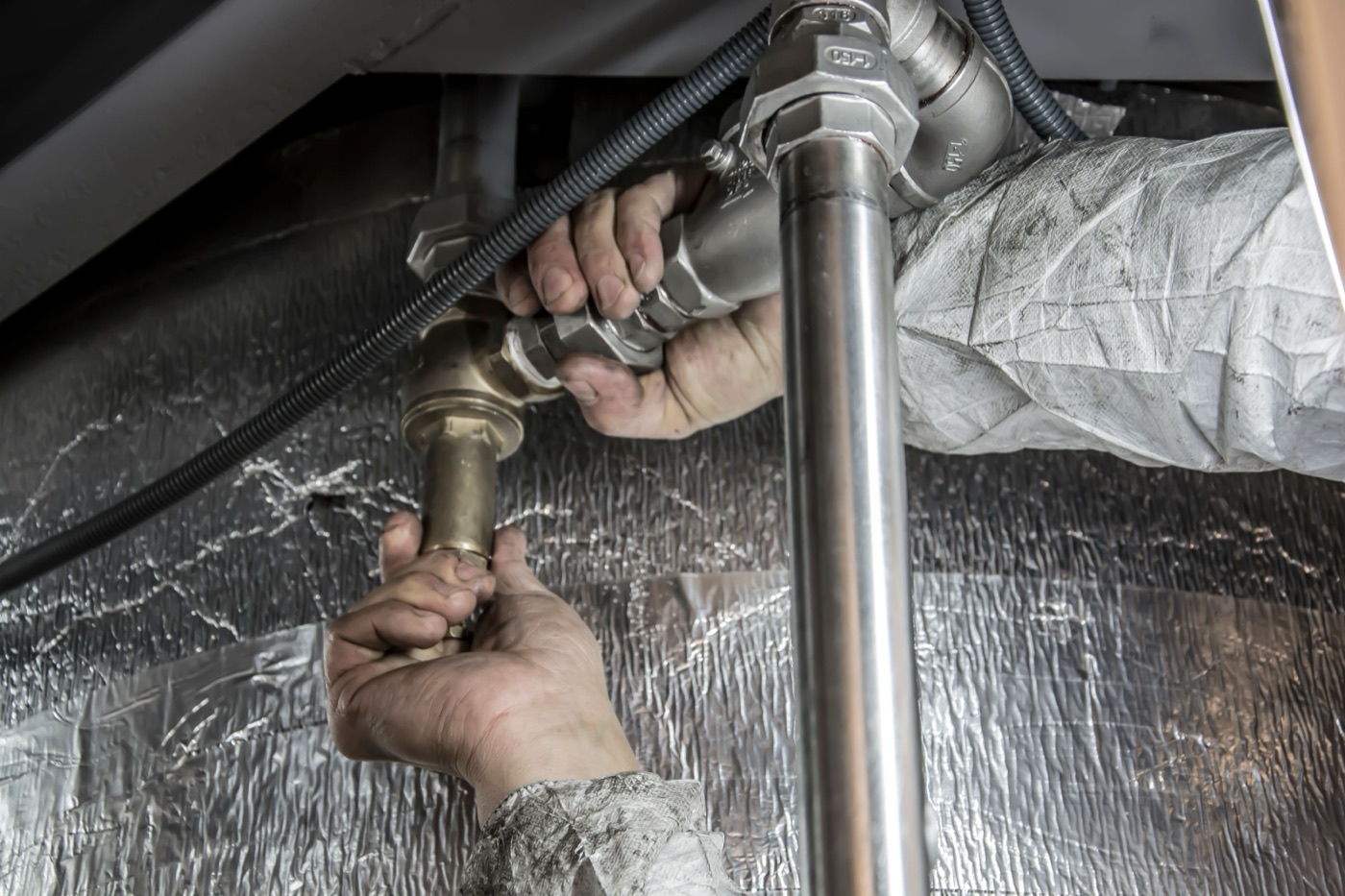 Close-up of a plumber's hands tightening brass and stainless steel pipe fittings against foil-faced insulation.