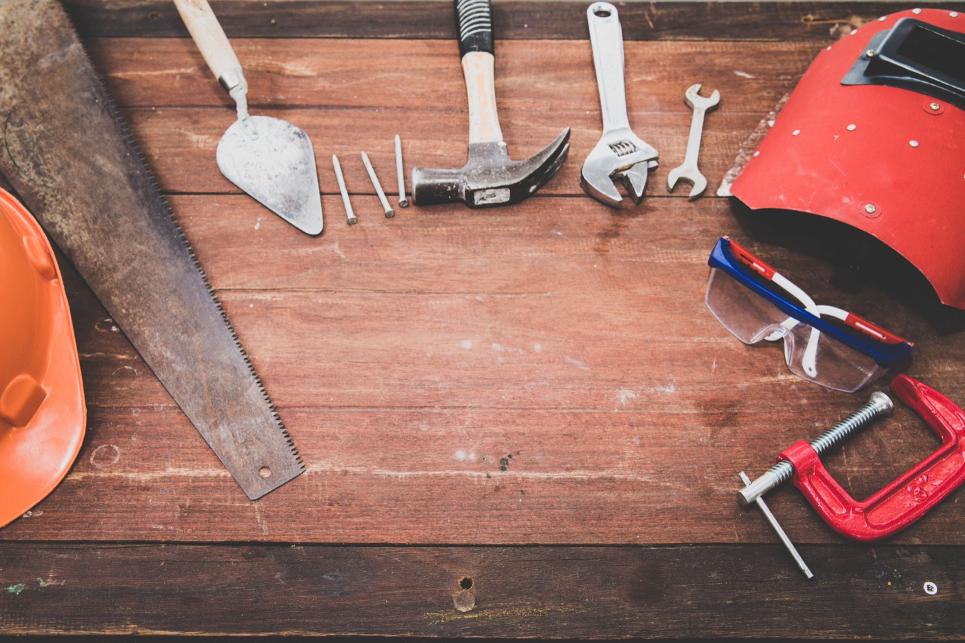 Overhead view of hand tools on a worn wooden workbench: a handsaw, trowel, claw hammer, nails, wrenches, safety glasses, a red C-clamp, an orange hard hat and a welding mask.