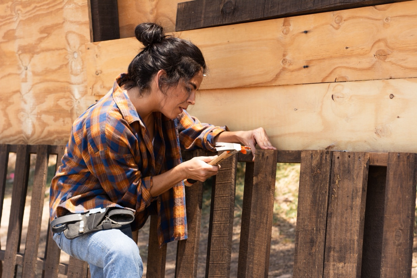 A woman in an orange and blue plaid flannel shirt and a tool belt kneels to hammer a nail into a weathered wooden fence board.